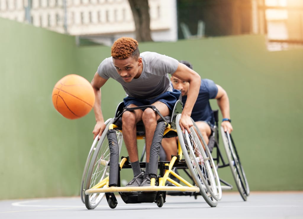 Two athletes in wheelchairs playing basketball on an outdoor court, one reaching for the ball.