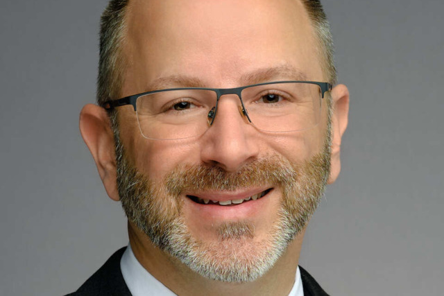 A person in a suit and pink tie smiling against a gray background.