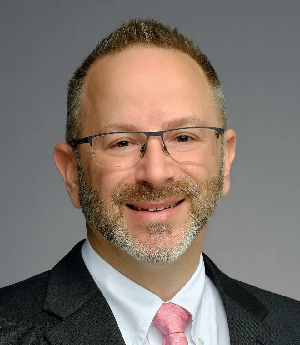 A person in a suit and pink tie smiling against a gray background.