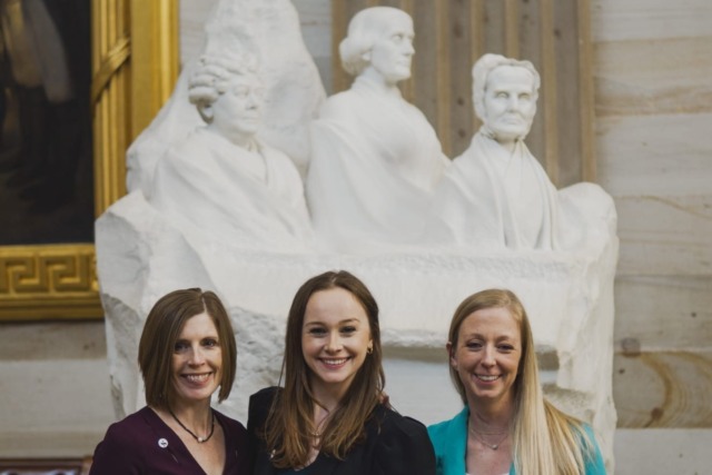 Three women standing in front of a white marble sculpture with busts of historical figures in a grand room.
