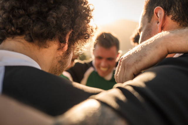 Rugby players in a huddle sharing a moment of teamwork and camaraderie under the warm sunlight on a playing field.