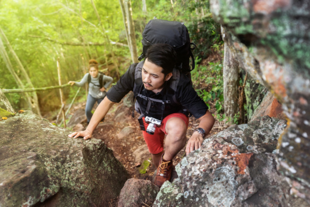 Two hikers climbing a rocky trail in a lush forest, one in focus with a backpack and camera, the other in the background.