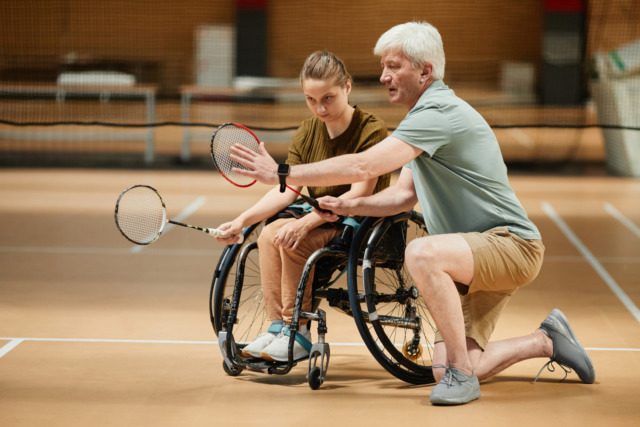 Instructor teaching wheelchair badminton to a young girl on a court.