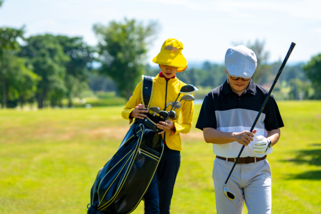 Two golfers walking on a sunny course, one carrying a golf bag and wearing a yellow outfit, the other adjusting a club.