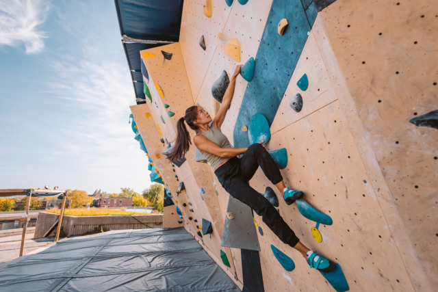 Woman climbing an outdoor bouldering wall on a sunny day, surrounded by colorful holds, wearing climbing shoes.