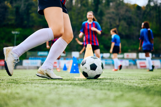 Soccer players practice dribbling around cones on a green field with teammates in the background during training session.