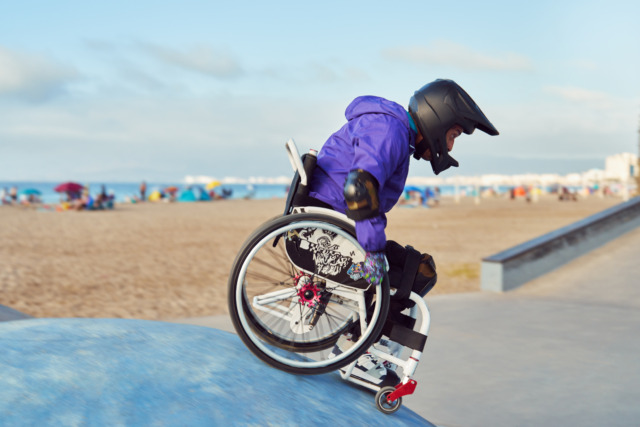 Person in a wheelchair wearing a helmet and protective gear at a skatepark by the beach.