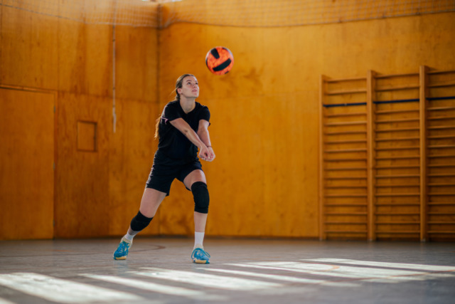 Female volleyball player in black jersey and knee pads bumping a ball indoors.