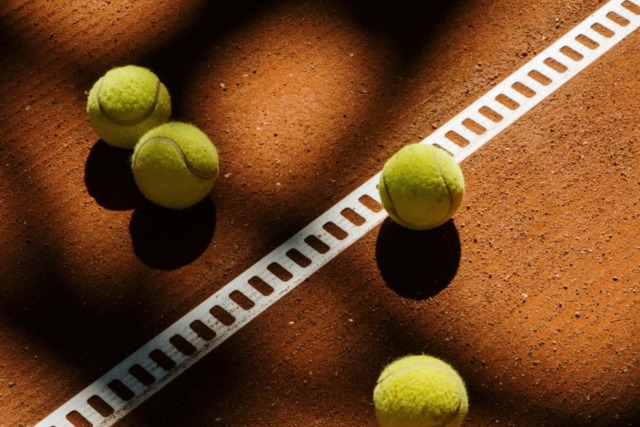 Tennis balls scattered on clay court with sunlight casting grid shadows over the scene.