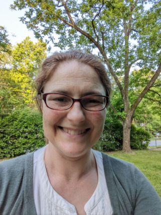A person with glasses and a smile stands outside in a green park with trees in the background.