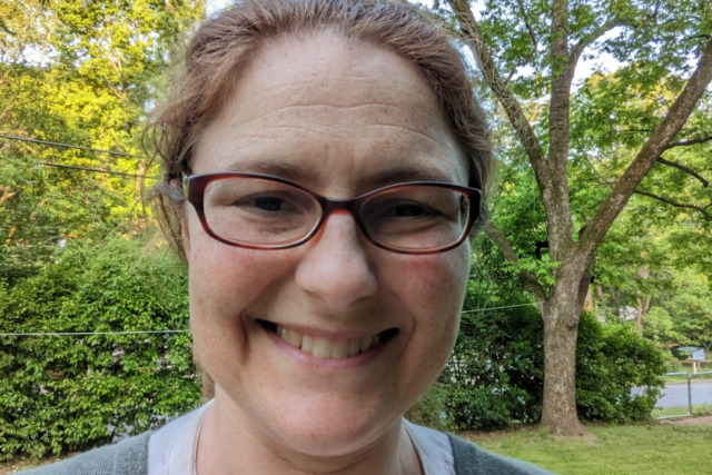 A person with glasses and a smile stands outside in a green park with trees in the background.