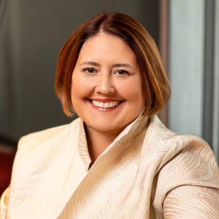 Smiling person wearing a textured light jacket, short auburn hair, sitting indoors facing the camera.