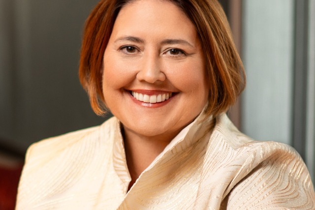 Smiling person wearing a textured light jacket, short auburn hair, sitting indoors facing the camera.
