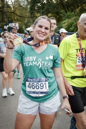 Woman smiling and holding a medal, wearing a green "Team Teal" shirt and race number 46691, after completing a marathon.