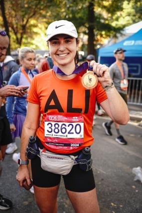 Runner smiling and holding a medal after a marathon, wearing an orange shirt and white cap, surrounded by other participants.