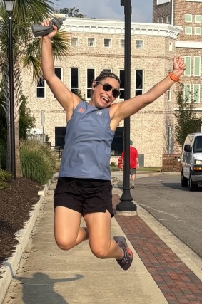 Person joyfully jumping on a sidewalk while holding a water bottle with buildings in the background.