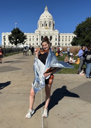 Woman in athletic gear wrapped in foil blanket, holding a medal in front of a large white capitol building on a sunny day.