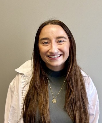 Smiling woman with long brown hair wearing a gray top and white jacket against a plain background.
