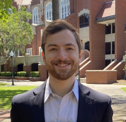 Smiling man in a dark suit stands outside a red-brick building with arched windows and greenery in the background.