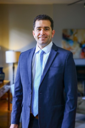 Man in a blue suit and light blue tie standing in a warmly lit room with a blurred background of a lamp and artwork.