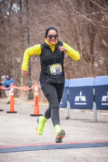 Runner crossing the finish line in a park race, wearing a yellow top and black vest, with a race bib and sunglasses.