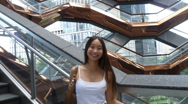Woman smiling in front of a complex staircase structure with copper-colored railings and cityscape background.