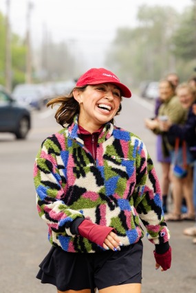 Person wearing colorful jacket and red cap, smiling and running on a street with blurred spectators in the background.