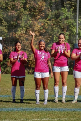 Five soccer players wearing pink jerseys standing on a field, one player waving, with trees in the background.