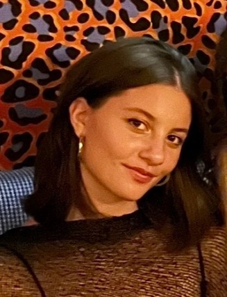 Person smiling with short dark hair, wearing a black top, standing in front of a patterned background.