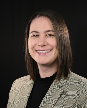 Smiling person with shoulder-length brown hair wearing a plaid blazer and black shirt against a black background.