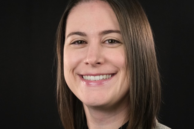 Smiling person with shoulder-length brown hair wearing a plaid blazer and black shirt against a black background.