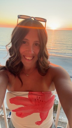 Woman smiling in front of a beach sunset, wearing sunglasses and a strapless top with a floral design.