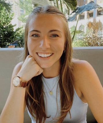 Person smiling while seated outdoors near greenery.