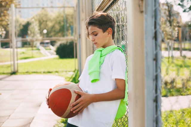 Boy holding basketball, leaning against a fence on a sunny day at an outdoor court.