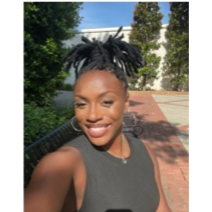 Smiling person with braided hair poses outdoors in a sunny setting with trees and a brick pathway in the background.