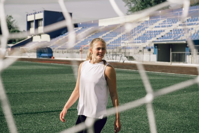 Soccer player in white tank top on field, with stadium seating in background, viewed through goal net.