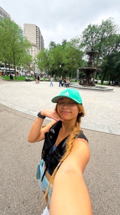 Person taking a selfie in a park with a fountain, wearing a green cap, trees and buildings in the background.