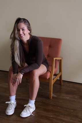 Person sitting on a chair, wearing casual attire with long hair and sneakers, smiling at the camera indoors.