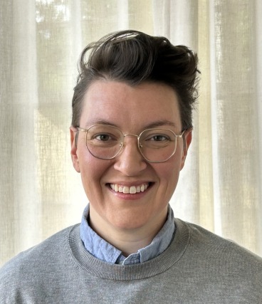 Person with short hair and glasses wearing a gray sweater over a light blue shirt, smiling in front of a beige curtain.
