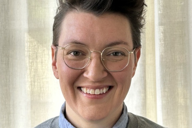 Person with short hair and glasses wearing a gray sweater over a light blue shirt, smiling in front of a beige curtain.