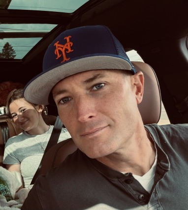 man in baseball cap taking car selfie with smiling woman in background