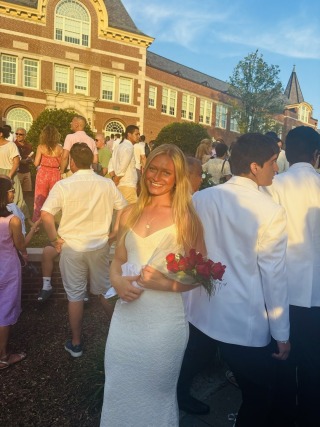 Woman in white dress holding flowers at an outdoor formal event with people and a large brick building in the background.