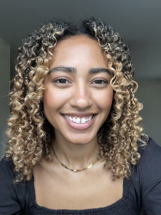 Smiling person with curly hair, wearing a black top and gold necklace, posed against a neutral background.