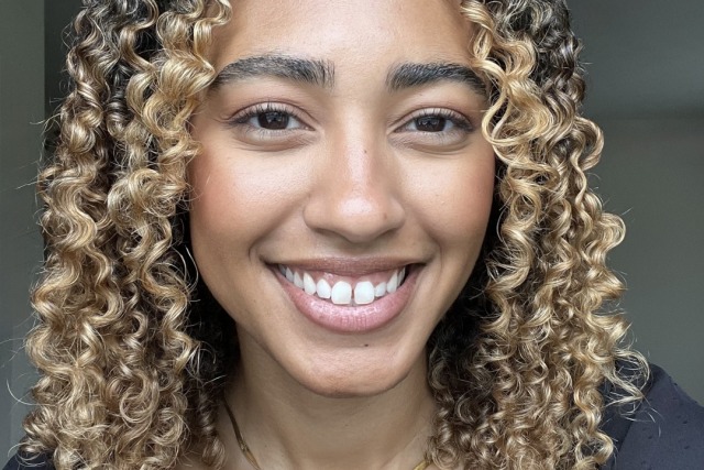 Smiling person with curly hair, wearing a black top and gold necklace, posed against a neutral background.