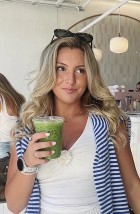 Person with long hair holding a cup of green iced beverage, standing inside a modern café.