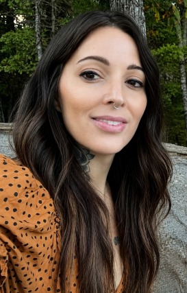 Woman with long dark hair and a nose ring, wearing a patterned orange blouse, smiling outdoors against a forest backdrop.